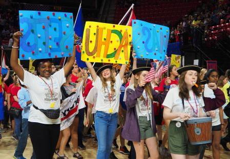 学生在全国比赛的NHD游行中行进.  学生们穿着NHD马萨诸塞州的t恤，戴着三角帽.  前排的三个学生举着牌子，上面写着 